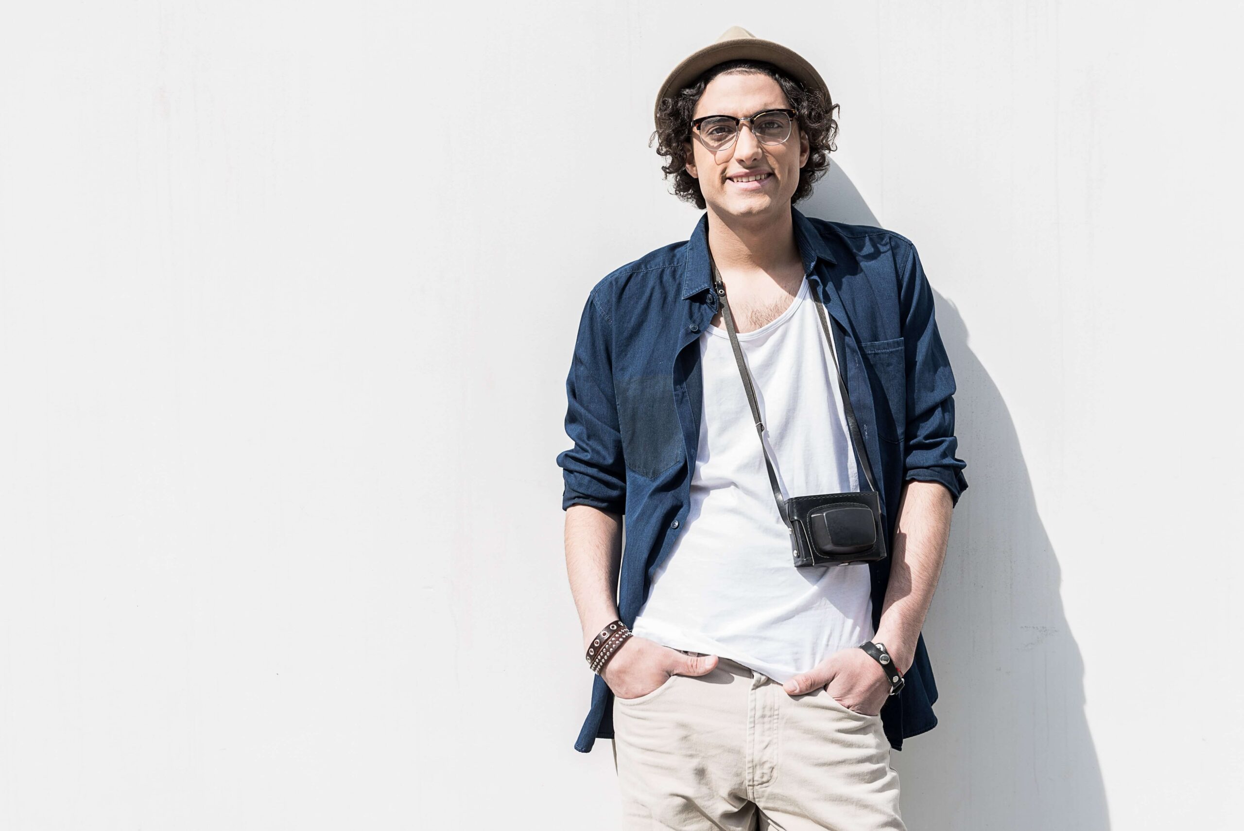 Positive vibes. Portrait of cheerful young man is leaning on white wall. His hands are in pockets of his pants. He is looking at camera with joy. Copy space in the left side
